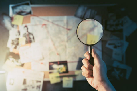 Detective board connecting clues with a man's hand holding a magnifying glass in the foreground
